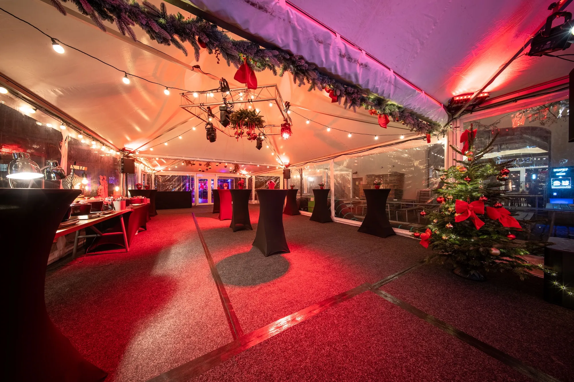 Vánočně vyzdobený stan s koktejlovými stolky, osvětlením a ozdobeným vánočním stromkem s červenými mašlemi.