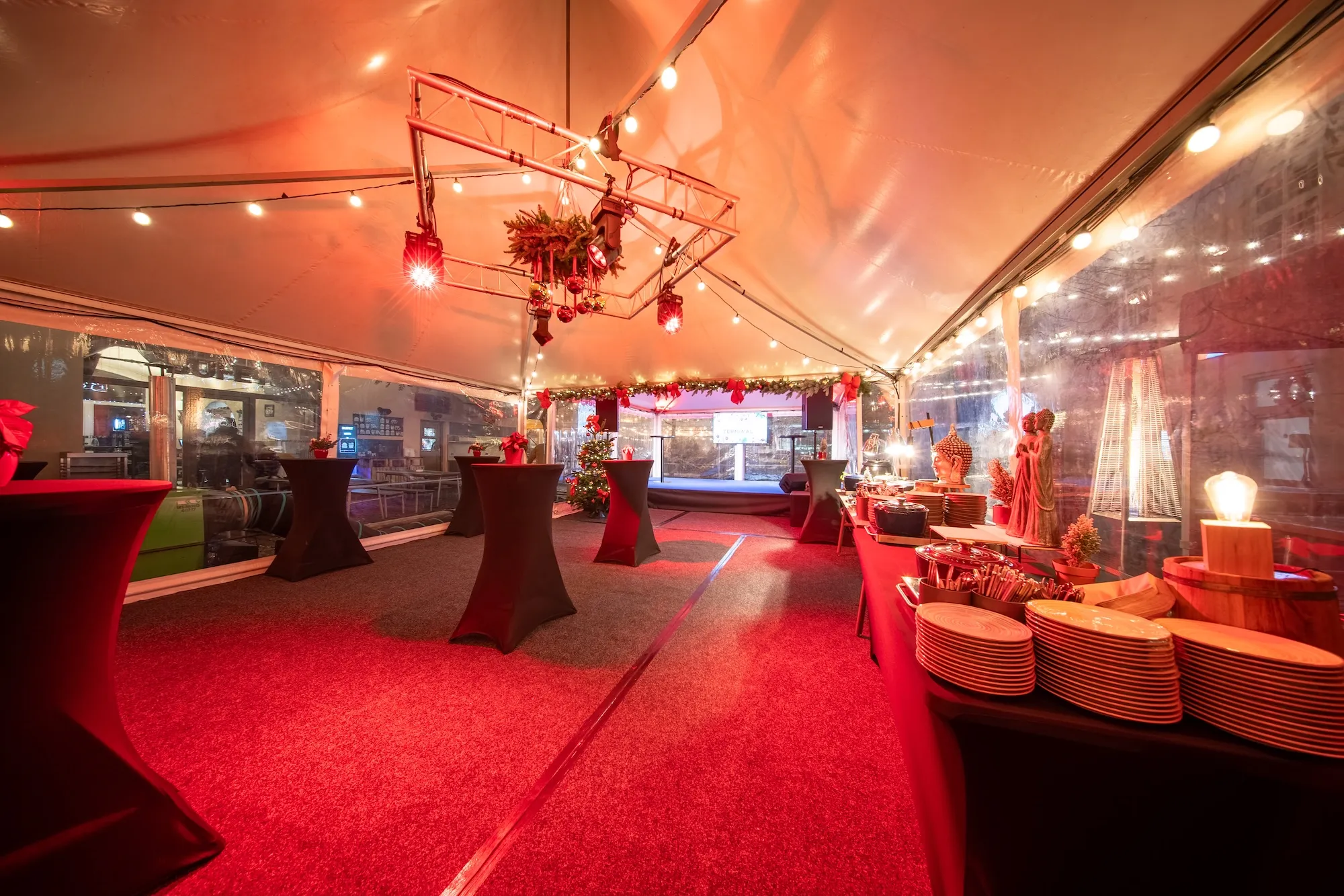 Vánočně vyzdobený prostor v bílé stanu s vysokými stojany, ozdobeným vánočním stromkem a stolem s talíři a dekoracemi.