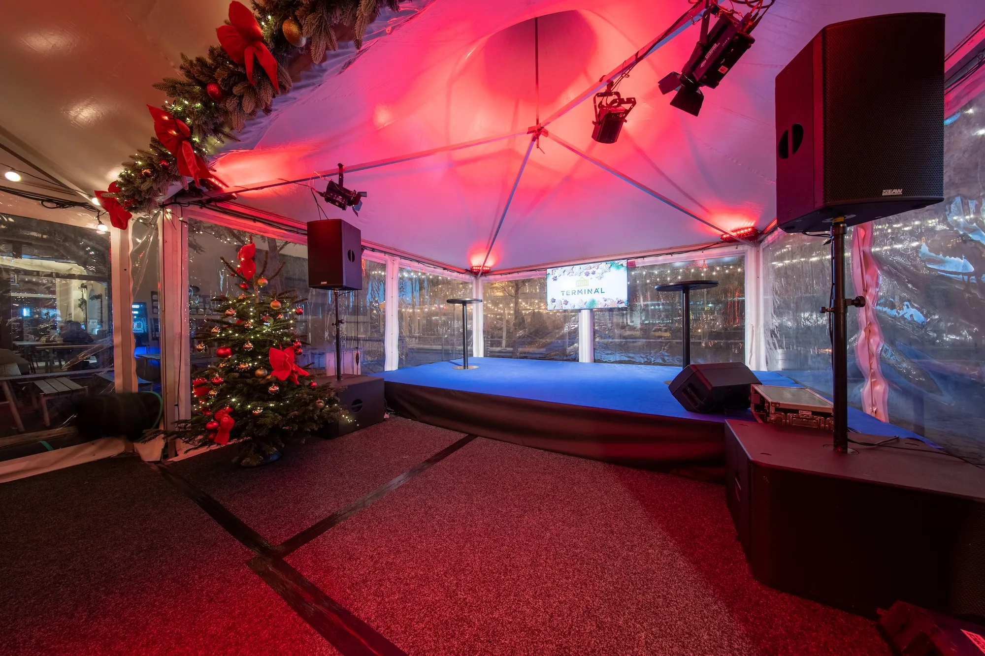 Vánočně vyzdobený pódium v jasně osvětleném stanu s vánočním stromkem se stužkami a světýlky.
