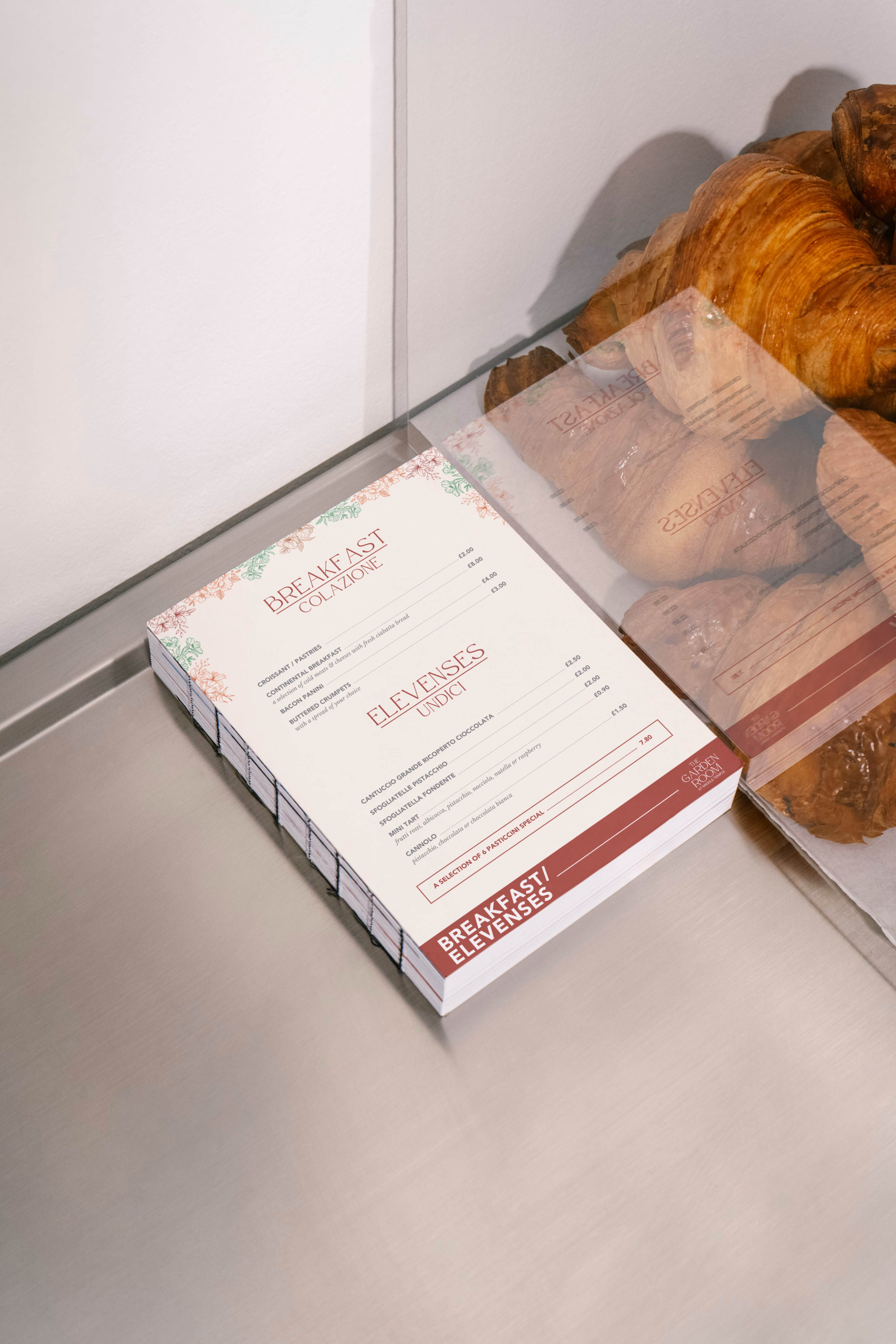 Stack of breakfast menus on a metal surface beside croissants under a glass cover.