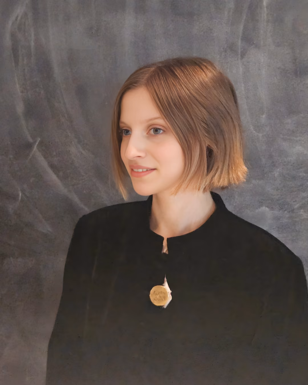 Young woman with short brown hair wearing a black top with a gold pendant, against a gray background.