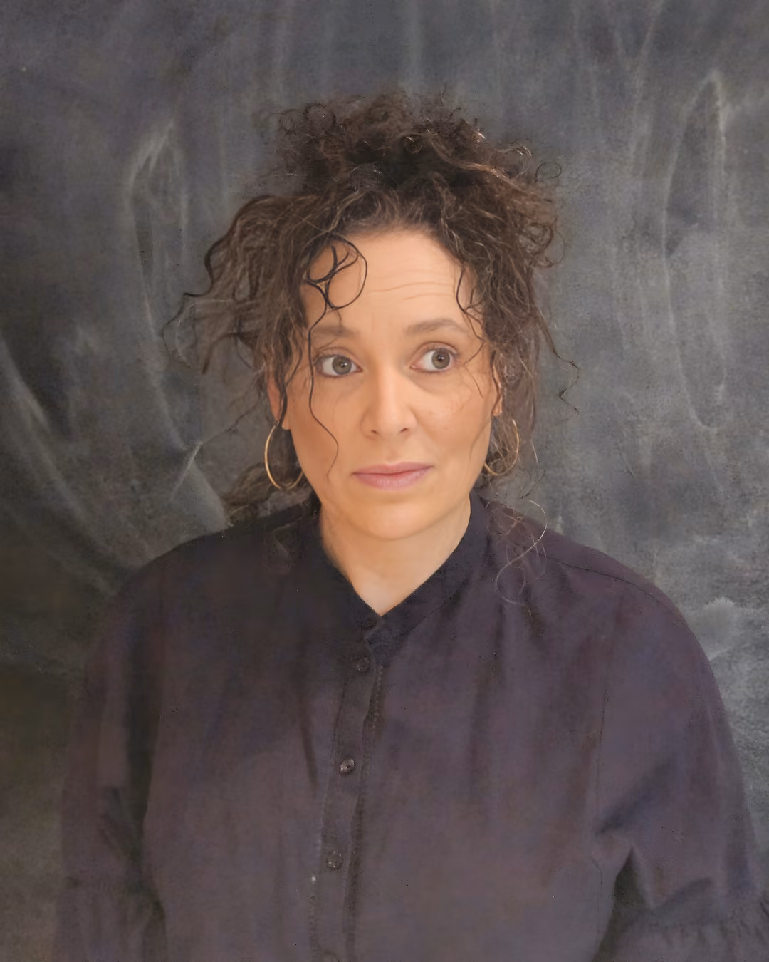 Woman with curly hair, hoop earrings, and a dark buttoned shirt looking to the side against a gray background.