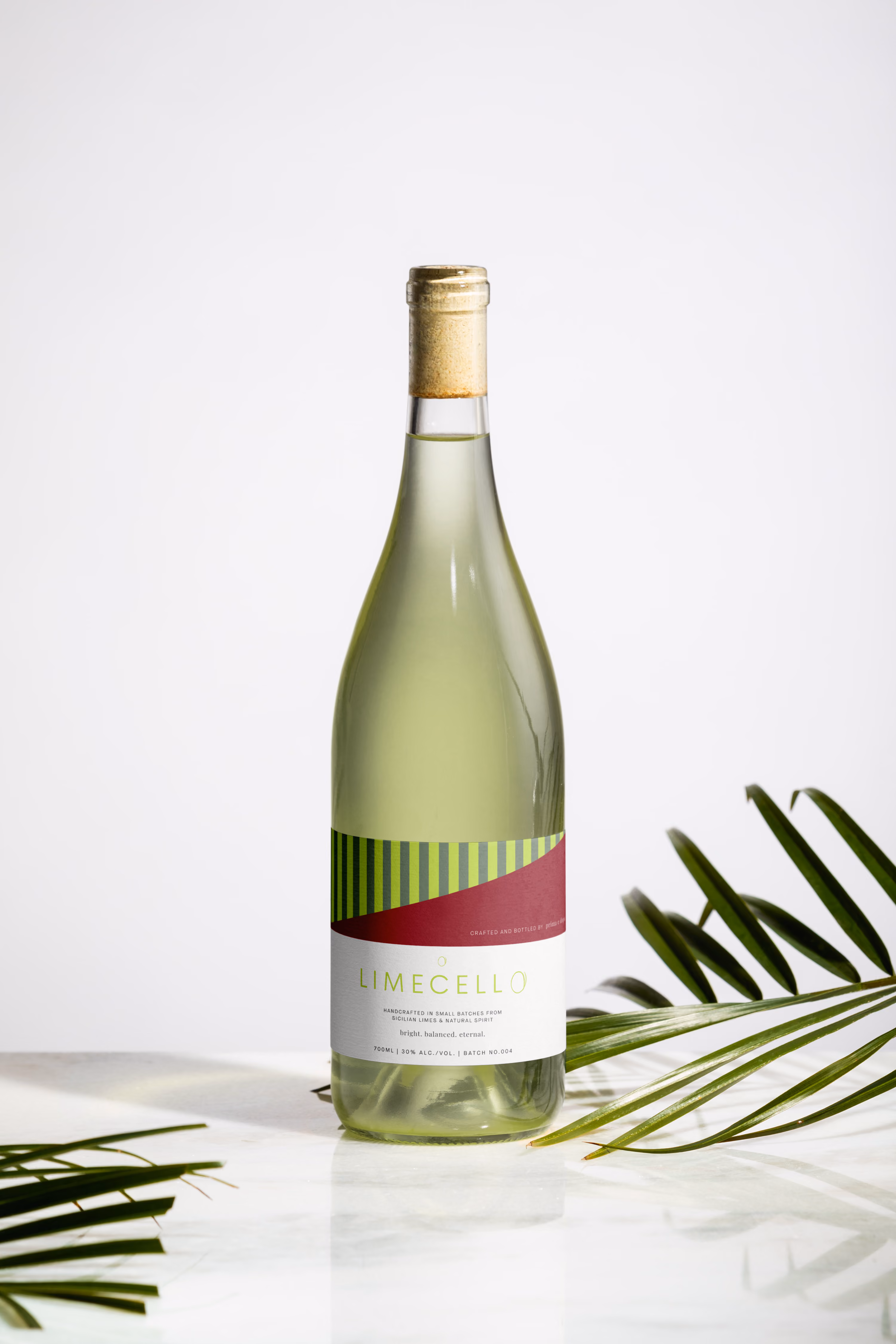 A green bottle of LIMECELLO liqueur with a red and green label on a white marble surface with palm leaves.