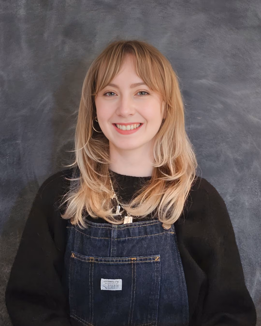 Smiling woman with blonde hair wearing a black sweater and denim overalls against a gray background.