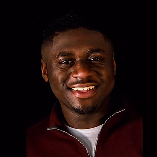 Portrait of a smiling young man against a black background wearing a dark jacket and white shirt.