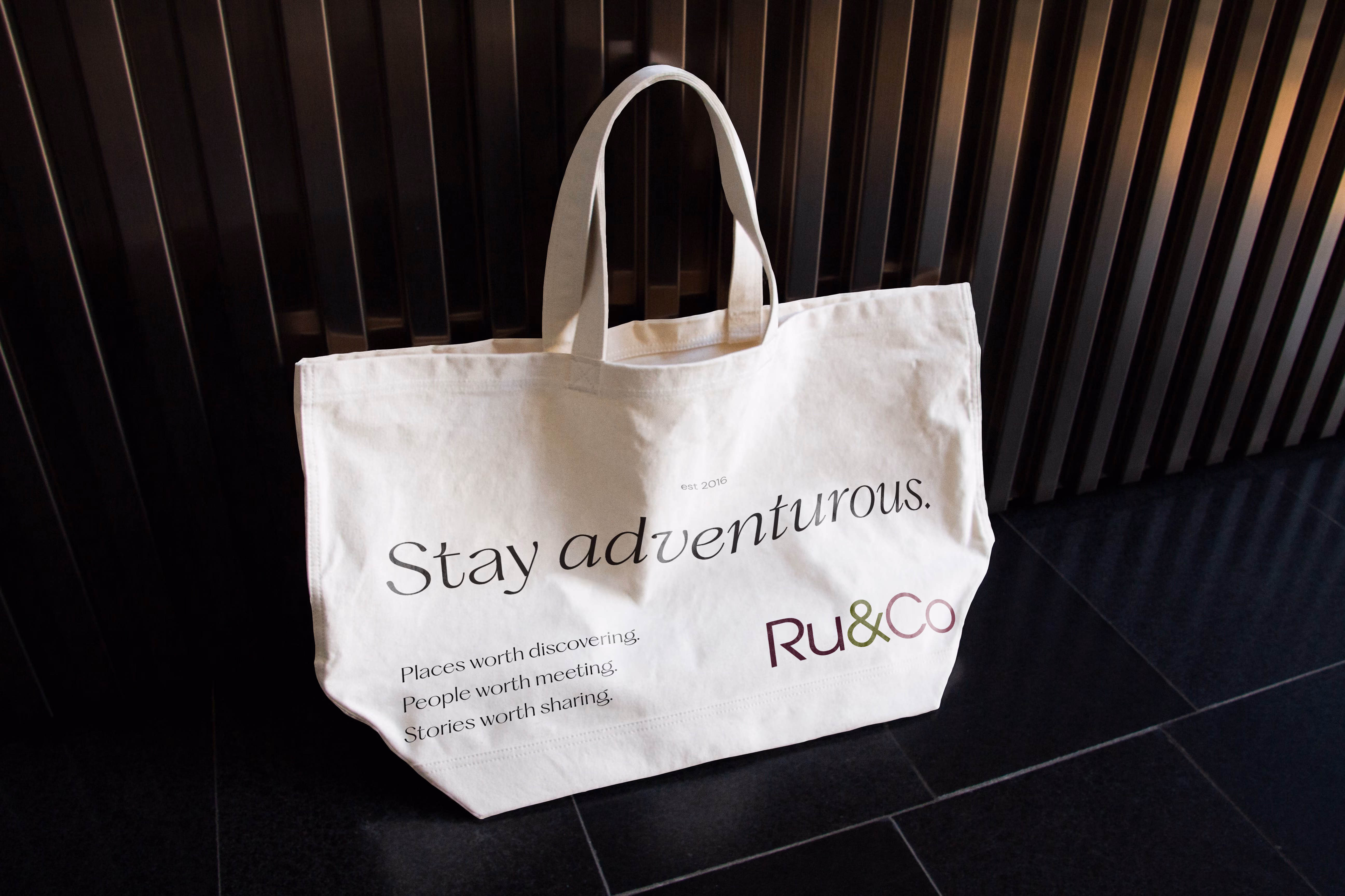White tote bag with text: Stay adventurous. Places worth discovering, people, stories worth sharing. Ru&Co logo.