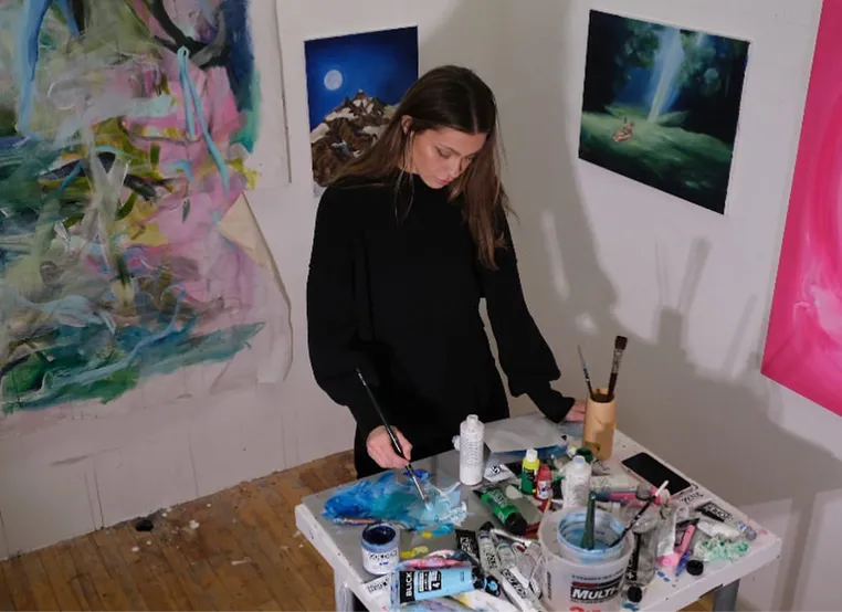 An artist in a black sweater paints at a cluttered table, surrounded by colorful paintings and various art supplies.