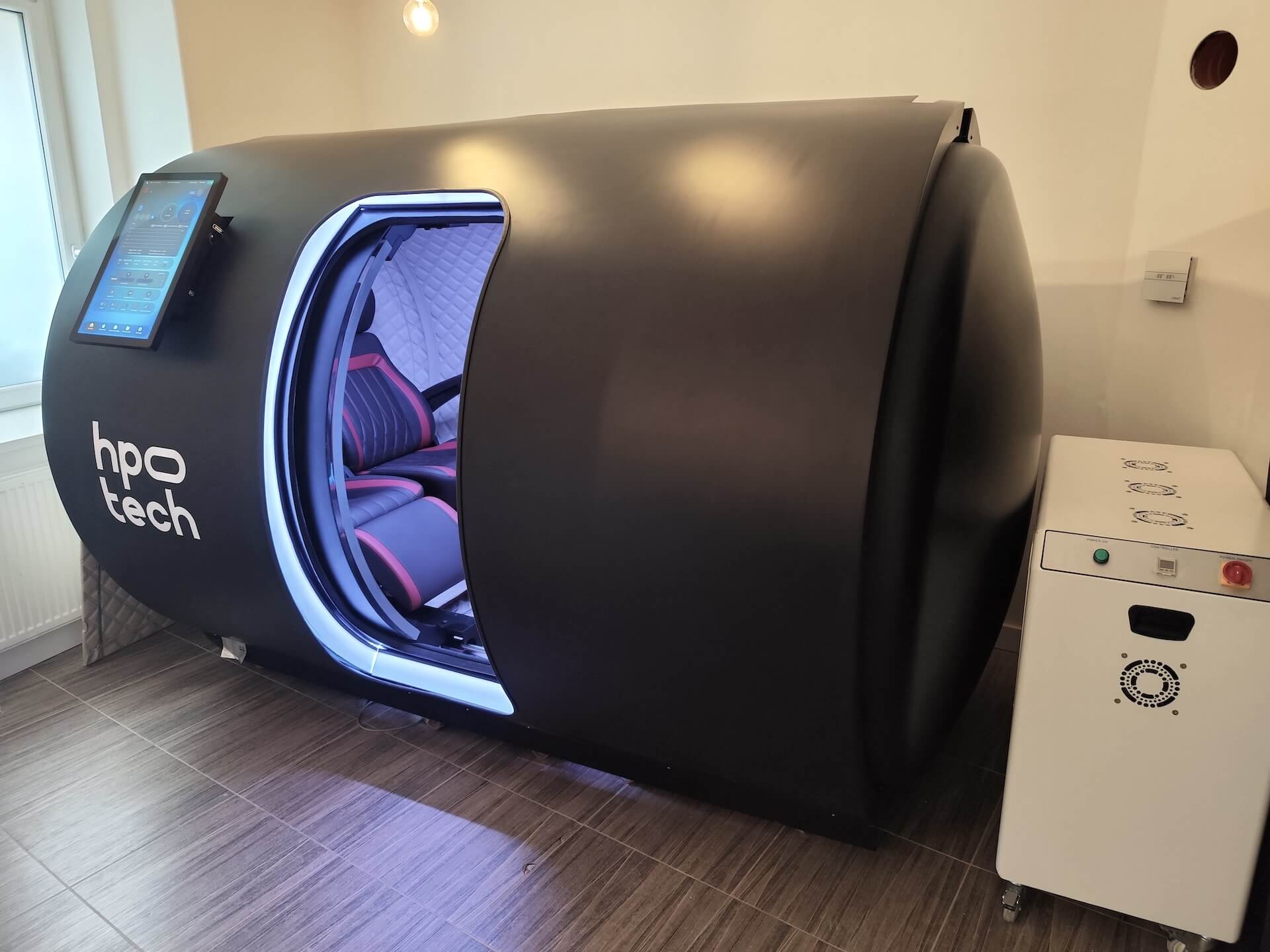 Černá kapsle krehabilitaci nebo relaxaci s otevřenými dveřmi a čalouněným sedadlem uvnitř, vedle ní bílá ovládací jednotka.