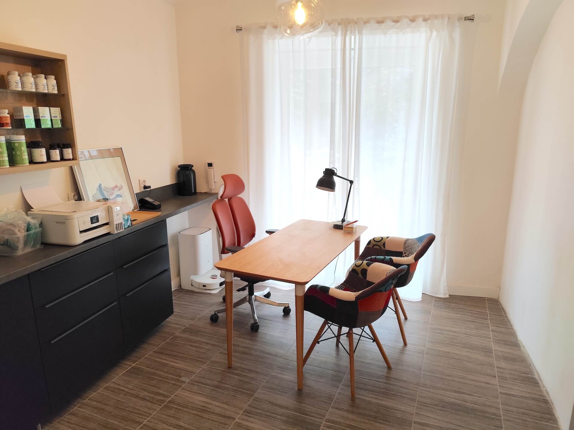 Moderní kancelář s dřevěným stolem, červenou kancelářskou židlí a dvěma barevnými křesly u okna se závěsem.