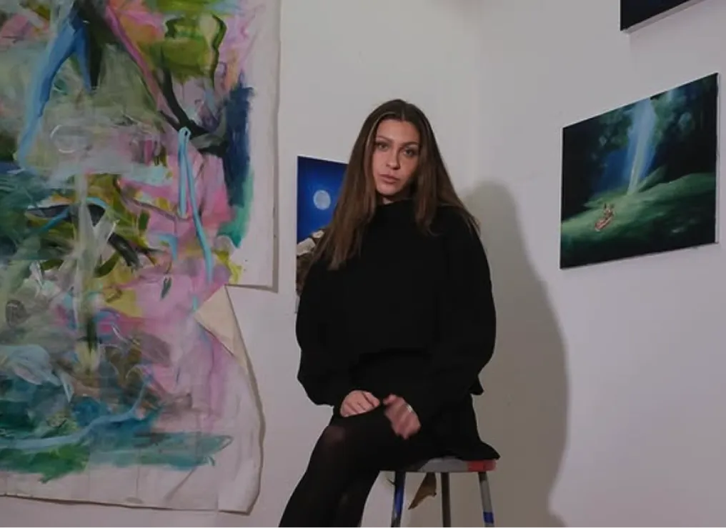 A person in black sits on a stool in a studio adorned with colorful abstracts and serene landscapes.