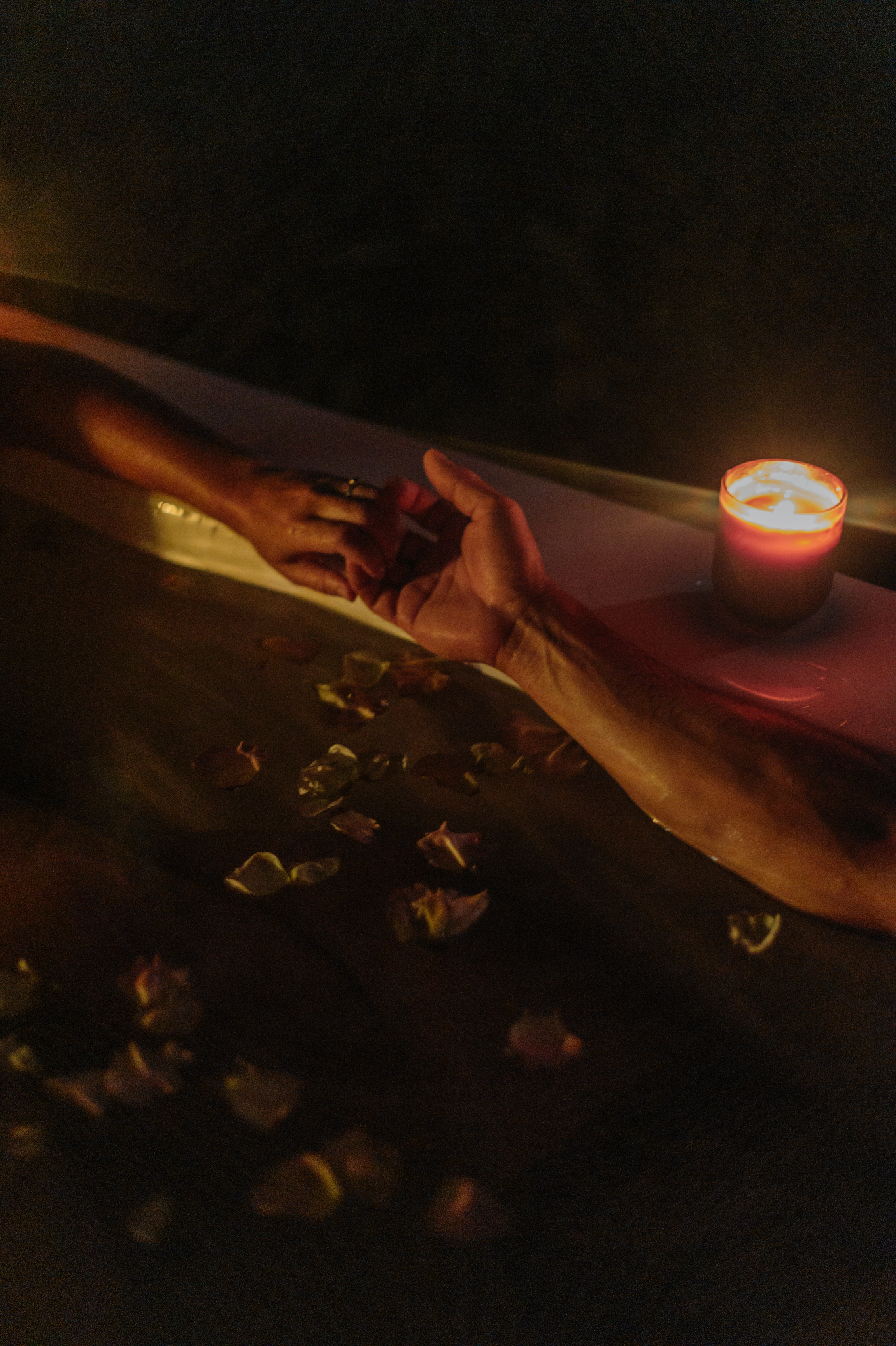 Two people holding hands across a candlelit outdoor bath at night, with rose petals floating on the warm water.