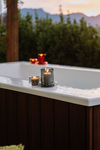 A timber-clad Star Baths outdoor bath at dusk, candles flickering along the edge, with native greenery and mountains beyond.