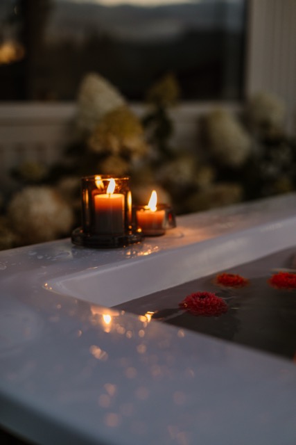 Close-up of an outdoor bath at dusk, lit candles on the rim and red dahlias floating on the water, white hydrangeas behind.