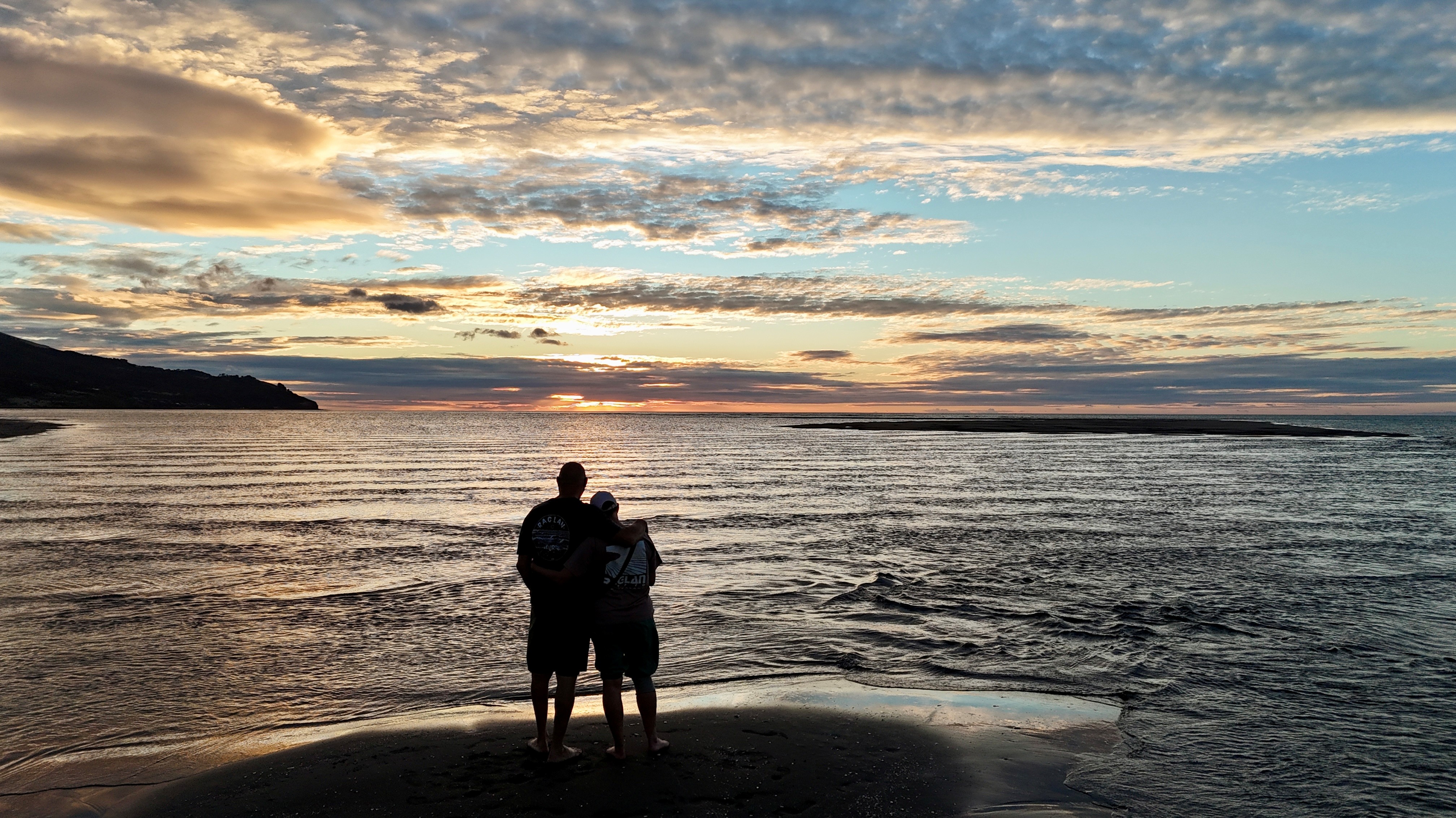 Ange and Anthony standing arm in arm on a Raglan beach at sunset, looking out over the low-tide sea.