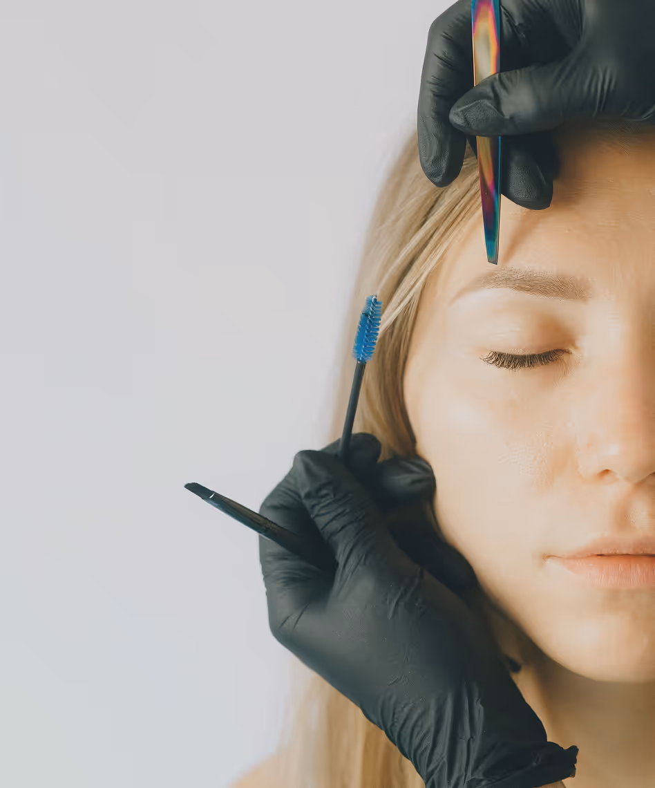 Close-up van vrouw met gesloten ogen die een wenkbrauwbehandeling krijgt van een specialist met zwarte handschoenen en pincet.