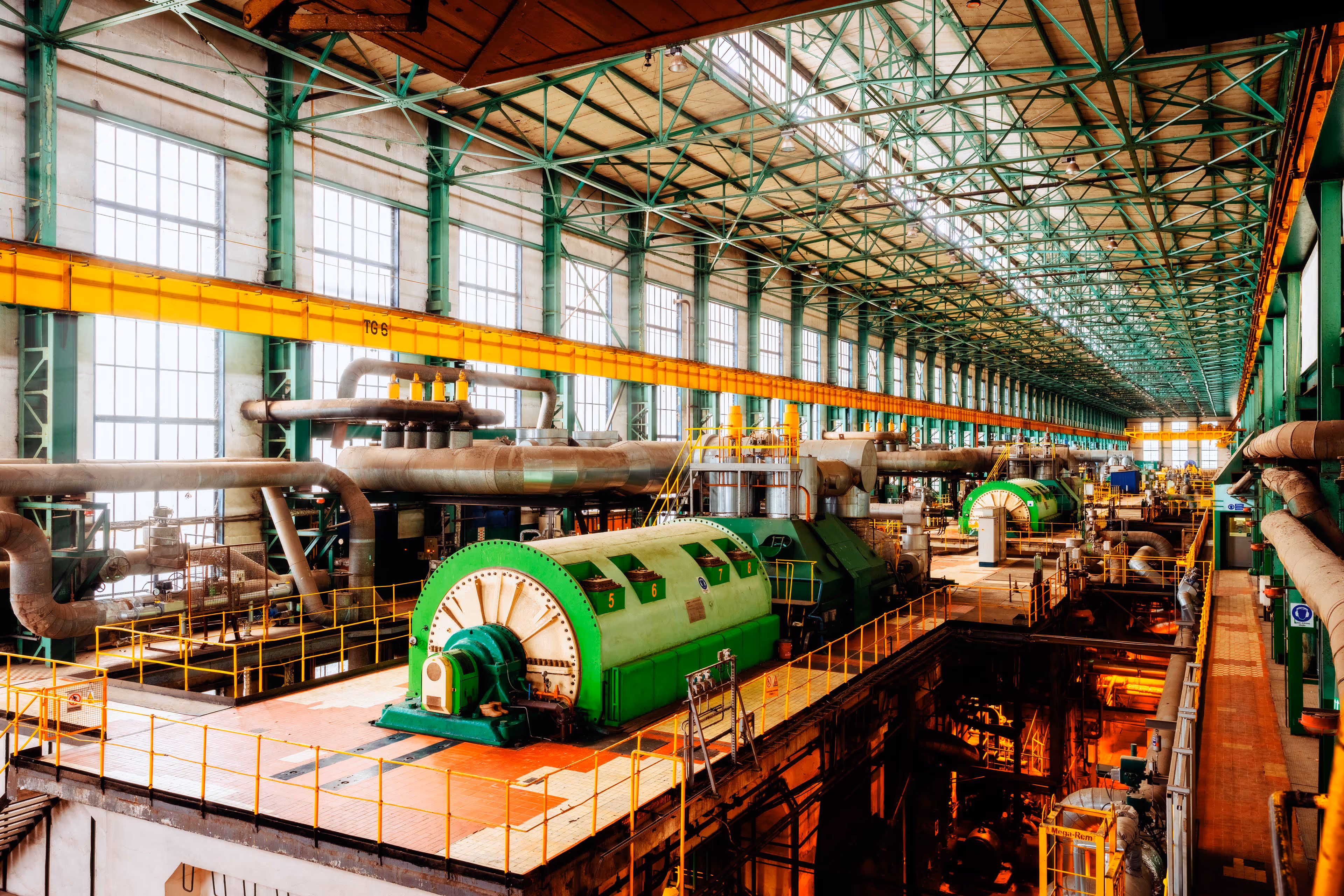 Interior of a vast industrial power plant hall featuring a series of large, green turbine generators mounted on raised platforms, each surrounded by yellow safety railings. The ceiling is supported by a grid of green steel beams and filled with rows of tall vertical windows that flood the space with natural light. Overhead runs a bright yellow gantry crane labeled TG-6, used for heavy maintenance. Massive grey pipelines weave across the facility, connecting machinery and infrastructure. Below the main level, glowing orange lighting reveals additional equipment and piping, highlighting the scale and complexity of energy production within the facility.