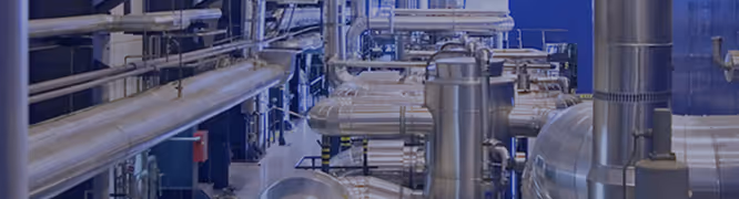 Interior view of a modern industrial facility filled with an intricate network of large stainless steel pipes, ducts, and cylindrical vessels. The pipes run horizontally and vertically, interconnected with joints and fittings, creating a dense mechanical environment. Some pipes are wrapped in insulation, and the entire space is brightly lit, reflecting off the polished metal surfaces. The image has a cool blue tint overlay, emphasizing a clean, high-tech, and precisely engineered atmosphere.