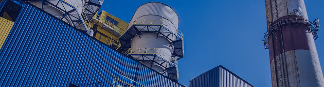 Wide-angle view of an industrial facility with large cylindrical structures at the center, likely silos or processing units, wrapped with metal platforms and yellow safety railings. The surrounding buildings are clad in blue corrugated metal panels, while a tall, weathered smokestack with red and white stripes rises on the right side. The sky is clear and vivid blue, and the entire image is lightly overlaid with a blue tint, giving the scene a clean, modern, and engineered appearance.