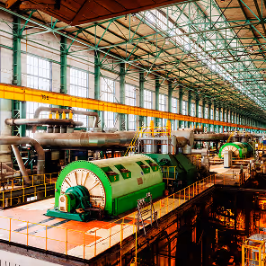Interior of a large industrial power plant with high ceilings and rows of tall windows along the walls. Dominating the space are several massive green turbine generators mounted on raised platforms, each enclosed in cylindrical housings with yellow railings for safety. Overhead, a green steel truss ceiling structure supports the building, and a yellow gantry crane runs along the top. The floor below the turbines is filled with large insulated pipes and additional equipment, all illuminated by warm lighting that highlights the scale and complexity of the facility.