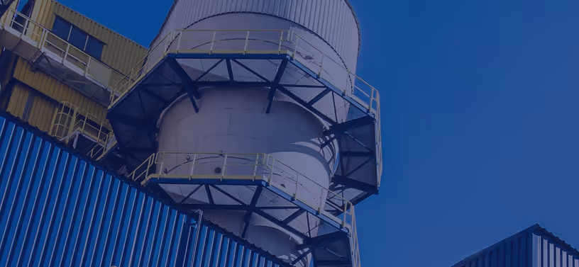 Close-up, low-angle view of a large cylindrical industrial structure made of metal, likely a storage silo or processing unit, surrounded by steel support platforms with yellow safety railings. The structure is connected to blue corrugated metal buildings and a yellow upper-level section with windows and access walkways. The sky in the background is clear and deep blue, and the entire image has a blue overlay that gives it a stylized, modern industrial appearance. The sharp lines and strong shadows emphasize the structure’s scale and engineering complexity.