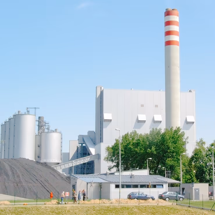 A wide view of a modern industrial facility featuring a large white building with rectangular vents, a tall red and white striped smokestack, and several cylindrical storage tanks on the left side. In front of the plant is a large mound of black coal or raw material, with a conveyor system leading toward the building. A small control office and several cars are visible near the base, along with a few people walking across a grassy area. Trees partially obscure the lower portion of the facility, and the sky is clear and blue, giving the scene a clean and organized appearance.