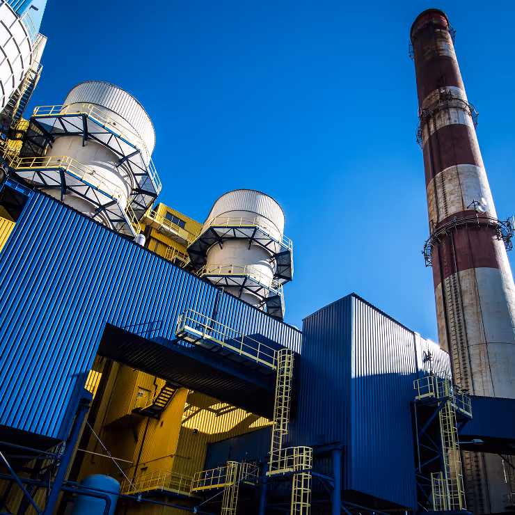 Low-angle view of a modern industrial facility featuring large cylindrical storage tanks with white ribbed surfaces and structural supports, corrugated blue and yellow metal buildings with walkways and ladders, and a tall, weathered smokestack with faded red and white stripes on the right side. The sky above is a deep, cloudless blue, and the entire scene is bathed in strong sunlight, casting sharp shadows and highlighting the geometric patterns and bold colors of the architecture. The image conveys a sense of scale, engineering precision, and industrial strength.