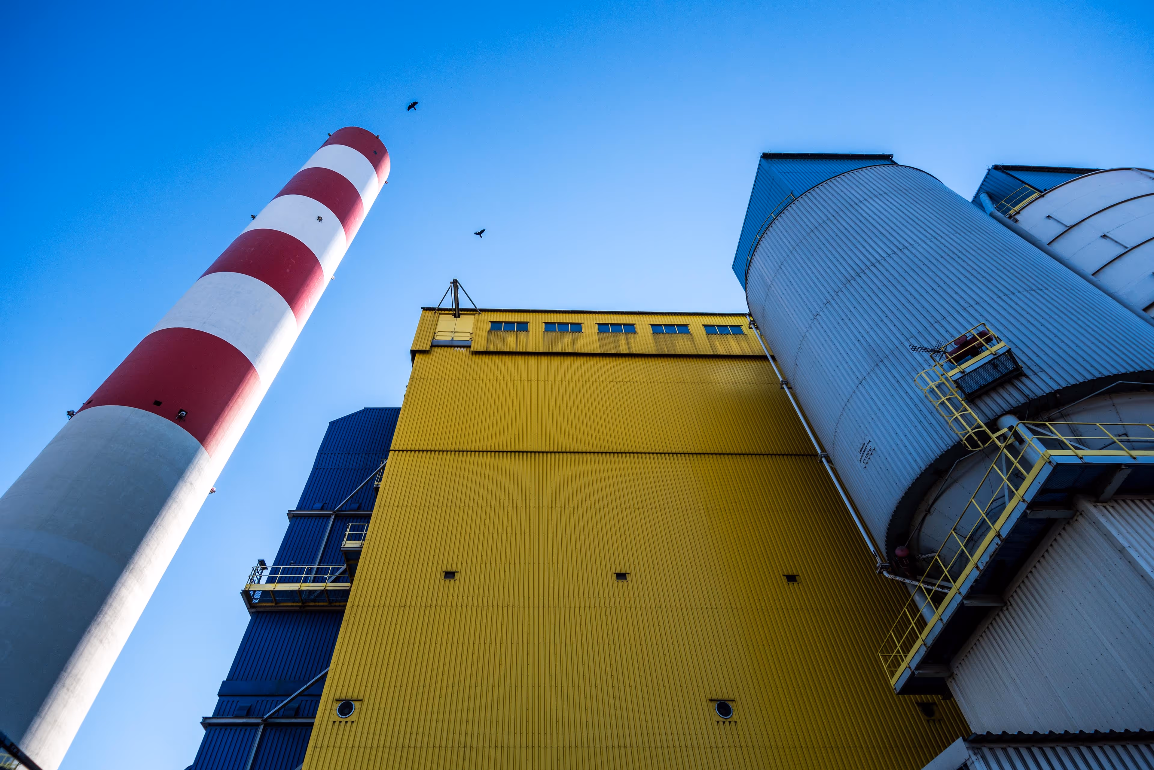 Low-angle view of an industrial facility featuring a tall red and white striped smokestack on the left, a large yellow corrugated metal building in the center, and cylindrical silver and blue storage tanks with yellow railings on the right. Additional blue structures and access platforms are visible on the left side of the building. The sky above is bright and cloudless, with three birds flying overhead, emphasizing the scale and verticality of the facility. The scene is clean, colorful, and sharply geometric, showcasing modern industrial architecture.