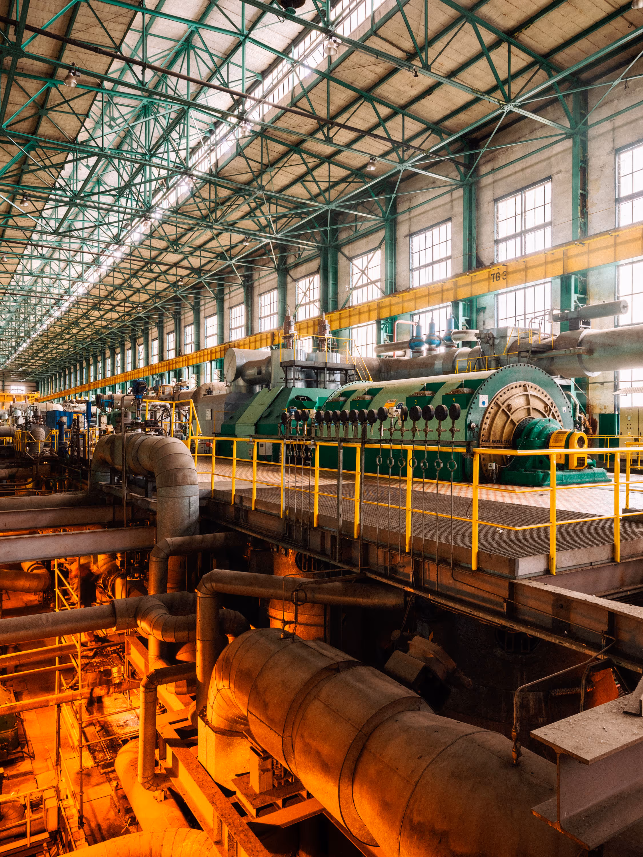 Interior of a large industrial power plant with a high ceiling supported by green steel beams and lined with tall windows that let in natural light. In the center, a massive green turbine generator with cylindrical housing and complex machinery is situated on a raised platform surrounded by yellow safety railings. Below the platform, numerous pipes and conduits run across the facility, illuminated by warm orange lighting, suggesting high-temperature operations. The environment is expansive, highly engineered, and meticulously maintained for heavy-duty energy production.