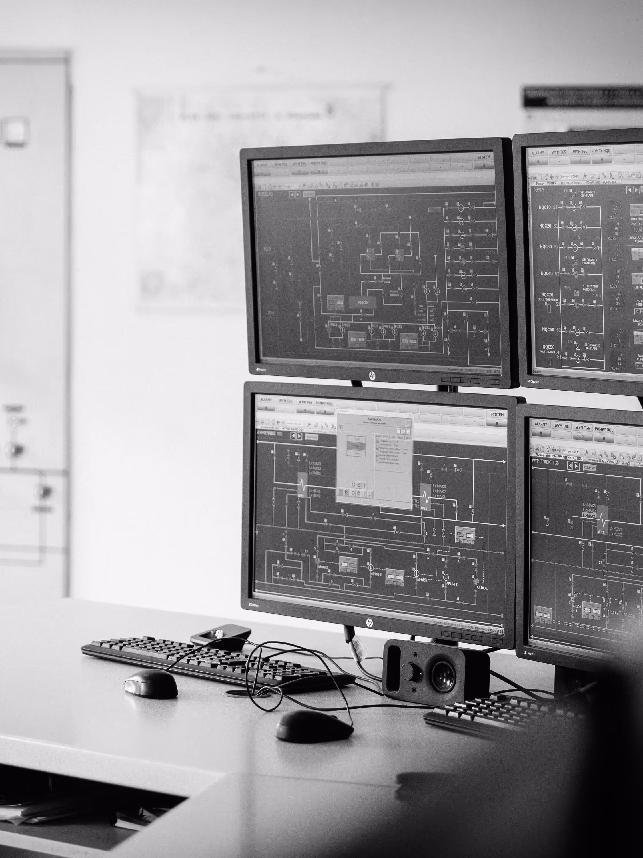 Black and white image of a workstation with four computer monitors arranged in a 2x2 grid, each displaying complex technical diagrams and system schematics, likely from industrial control software. The desk below holds two keyboards, two computer mice, a pair of eyeglasses, and various cables, suggesting active monitoring or configuration work. The background is out of focus but shows part of an office or control room wall. The scene reflects a high-tech, data-driven environment used for managing or supervising industrial processes.