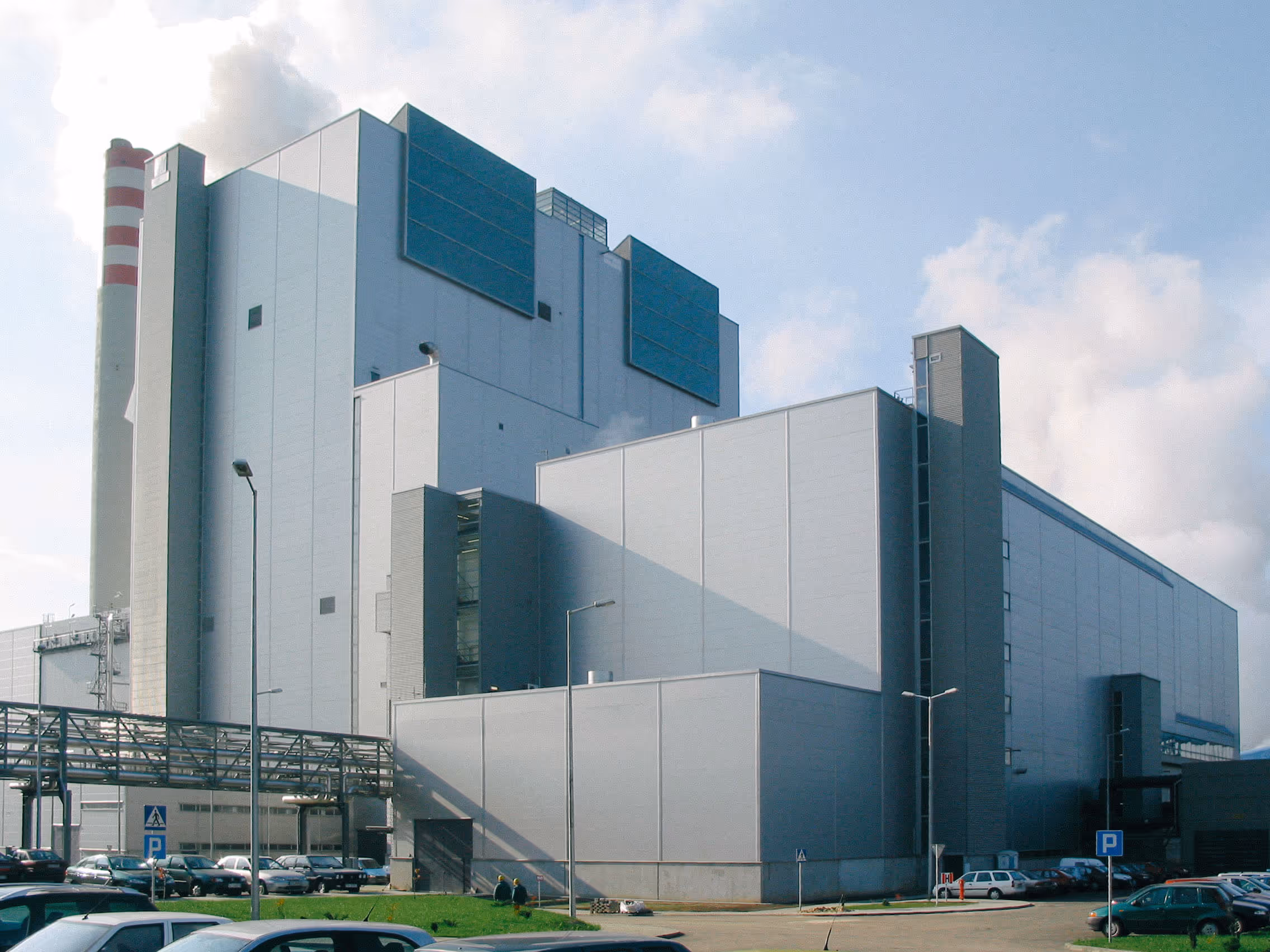 A large modern industrial facility composed of interconnected rectangular buildings with smooth, light grey metal panel exteriors. The structure rises in stepped levels with minimal windows, except for a few small square ones and some large vent-like panels painted in dark teal. The complex is surrounded by a clean, paved area with parked cars and streetlights. Steam or vapor can be seen rising from the top, indicating active industrial processes. The sky is partly cloudy, and the scene is brightly lit by daylight, highlighting the clean and functional architecture.