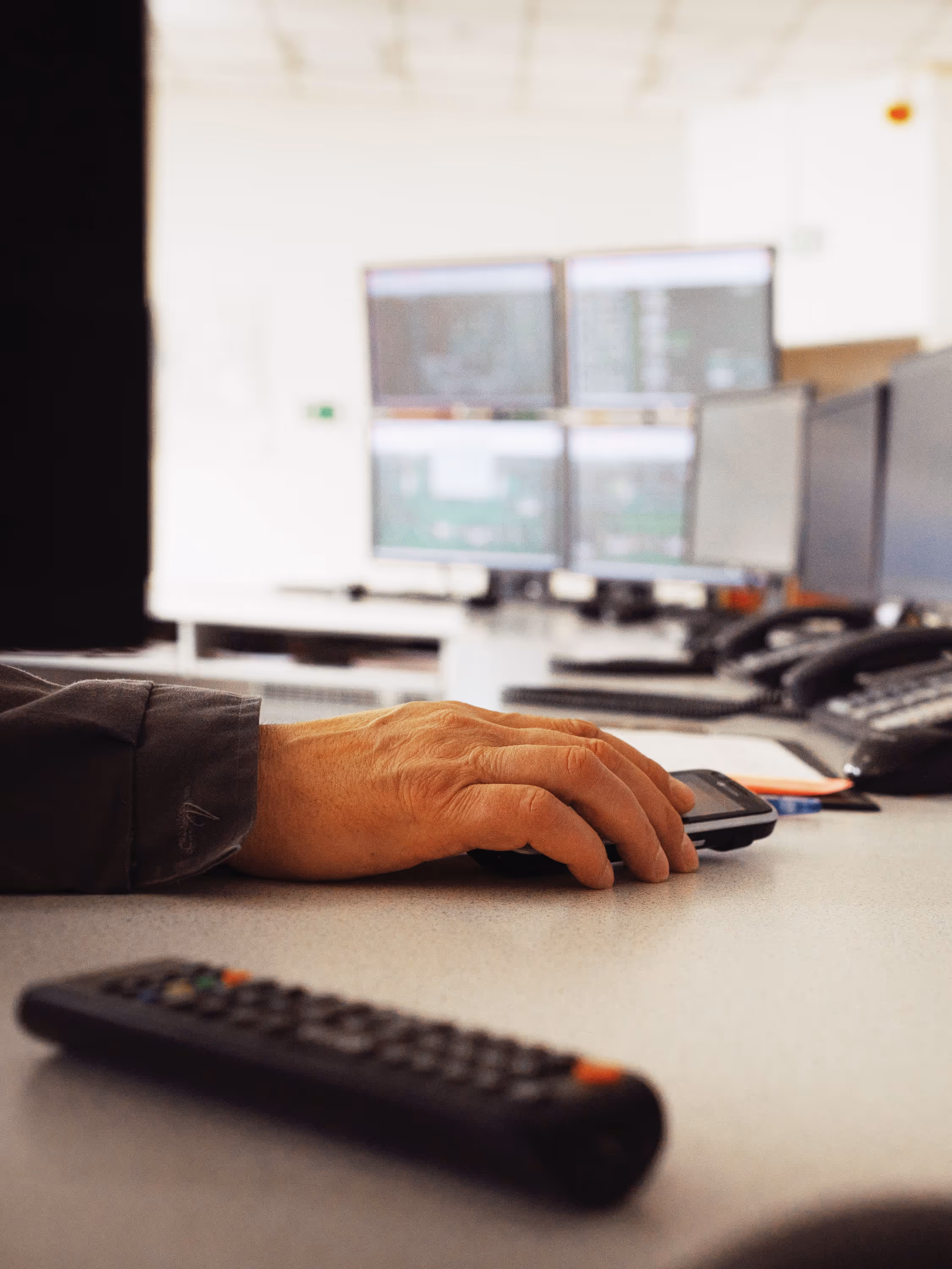 A close-up view of a person’s hand operating a computer mouse on a white desk in a modern control room. In the background, several large computer monitors display data and graphical interfaces, likely used for monitoring industrial systems or processes. The person is wearing a dark jacket, and other equipment such as telephones and a remote control are visible on the desk. The lighting is bright, emphasizing the high-tech and organized environment.