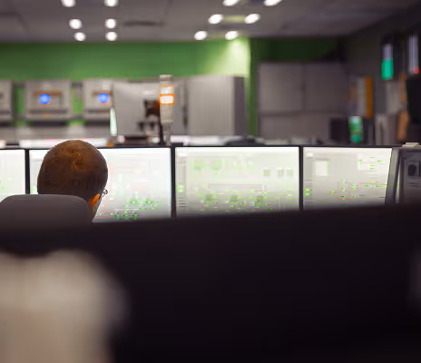 A man seated in a modern control room, viewed from behind as he monitors multiple computer screens filled with bright, data-rich system diagrams. The room is softly lit with ceiling lights, and the walls are painted green. In the background, several control panels with glowing blue indicators are mounted on a wall, and a vertical signal light with orange and white indicators stands atop a workstation. The atmosphere is quiet and focused, suggesting real-time monitoring of industrial or utility systems.