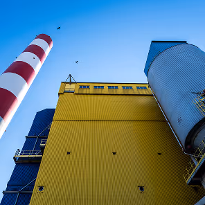 ChatGPT řekl:
Low-angle view of an industrial complex featuring a tall red and white striped smokestack on the left, a massive yellow corrugated building in the center, and a cylindrical silver-blue silo structure on the right. Additional blue structures with industrial walkways and platforms are visible on the left. Three birds fly high in the clear blue sky, adding a sense of scale and contrast to the bold architectural lines and vivid colors of the facility. The image conveys the scale and precision of heavy industry.