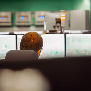 A man viewed from behind sitting in a control room, closely monitoring several computer screens displaying complex data and system diagrams. The screens cast a soft glow on his face as he focuses intently. In the background, a green wall and a row of equipment cabinets with blue-lit indicators can be seen, reinforcing the technical and high-security nature of the environment. A signal light with orange and white indicators is mounted on top of one workstation, suggesting an active industrial or utility monitoring setup.