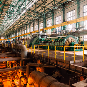 Interior of a large industrial power plant with a high ceiling supported by green steel beams and lined with tall windows that let in natural light. In the center, a massive green turbine generator with cylindrical housing and complex machinery is situated on a raised platform surrounded by yellow safety railings. Below the platform, numerous pipes and conduits run across the facility, illuminated by warm orange lighting, suggesting high-temperature operations. The environment is expansive, highly engineered, and meticulously maintained for heavy-duty energy production.