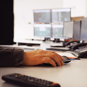 A close-up view of a person’s hand operating a computer mouse on a white desk in a modern control room. In the background, several large computer monitors display data and graphical interfaces, likely used for monitoring industrial systems or processes. The person is wearing a dark jacket, and other equipment such as telephones and a remote control are visible on the desk. The lighting is bright, emphasizing the high-tech and organized environment.