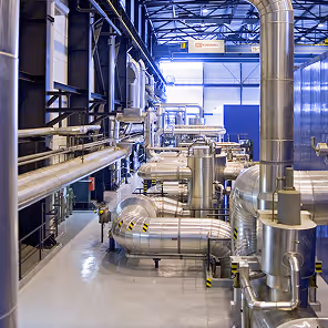 Interior of a high-tech industrial facility filled with large, shiny metal pipes and ducts running in various directions. The pipes are insulated and fitted with joints, valves, and black-and-yellow safety markings. The floor is clean and polished, and metal railings line the walkways for worker safety. The space is brightly lit with overhead industrial lighting, and a high ceiling supported by steel beams gives the room a spacious, organized appearance. The overall impression is of a modern, well-maintained energy or manufacturing plant.