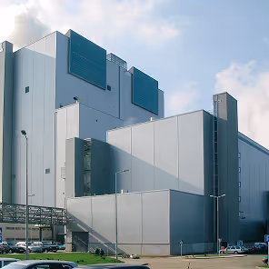 A large modern industrial facility composed of interconnected rectangular buildings with smooth, light grey metal panel exteriors. The structure rises in stepped levels with minimal windows, except for a few small square ones and some large vent-like panels painted in dark teal. The complex is surrounded by a clean, paved area with parked cars and streetlights. Steam or vapor can be seen rising from the top, indicating active industrial processes. The sky is partly cloudy, and the scene is brightly lit by daylight, highlighting the clean and functional architecture.