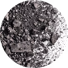 A close-up view of a pile of crushed rock or mineral fragments, captured in a circular frame. The surface is a mix of fine dust and variously sized sharp-edged pieces, ranging from tiny grains to larger, jagged chunks. The texture appears gritty and uneven, with a predominantly dark grey coloration. The fragments are scattered naturally, creating a raw, unrefined look that suggests industrial or geological processing.