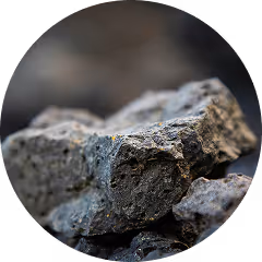 A close-up image of several dark grey chunks of coal or mineral rock, shown in sharp focus with a blurred background. The texture of the rock is rough and uneven, with small cracks, ridges, and a matte surface that catches light subtly. Tiny flecks of rust-colored particles are scattered on the surface, adding to the natural, earthy appearance. The image is framed in a circular crop, giving it a clean and contained presentation.
