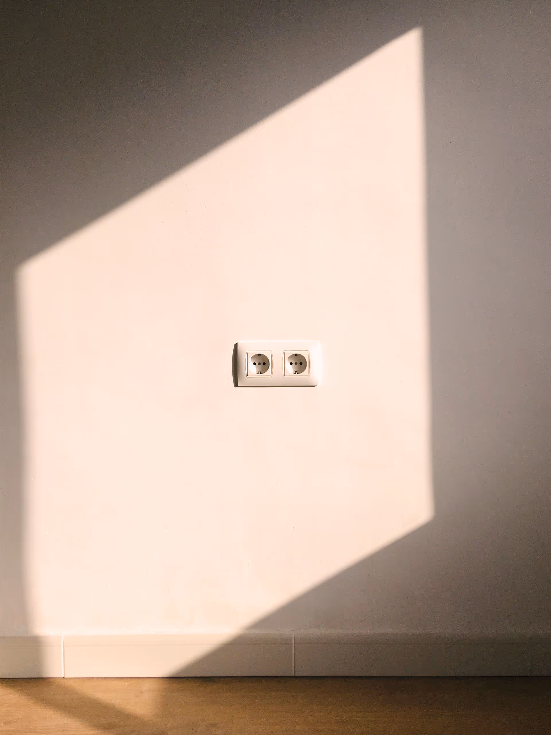 A minimalistic indoor scene showing a clean white wall with a double electrical outlet positioned slightly above a light-colored tile baseboard. A sharply defined trapezoidal shadow, cast by sunlight entering from an unseen window, covers part of the wall and floor, creating a strong contrast between light and dark areas. The wooden floor at the bottom adds warmth to the otherwise neutral setting. The image evokes a sense of calm and simplicity through its clean lines, natural lighting, and geometric composition.