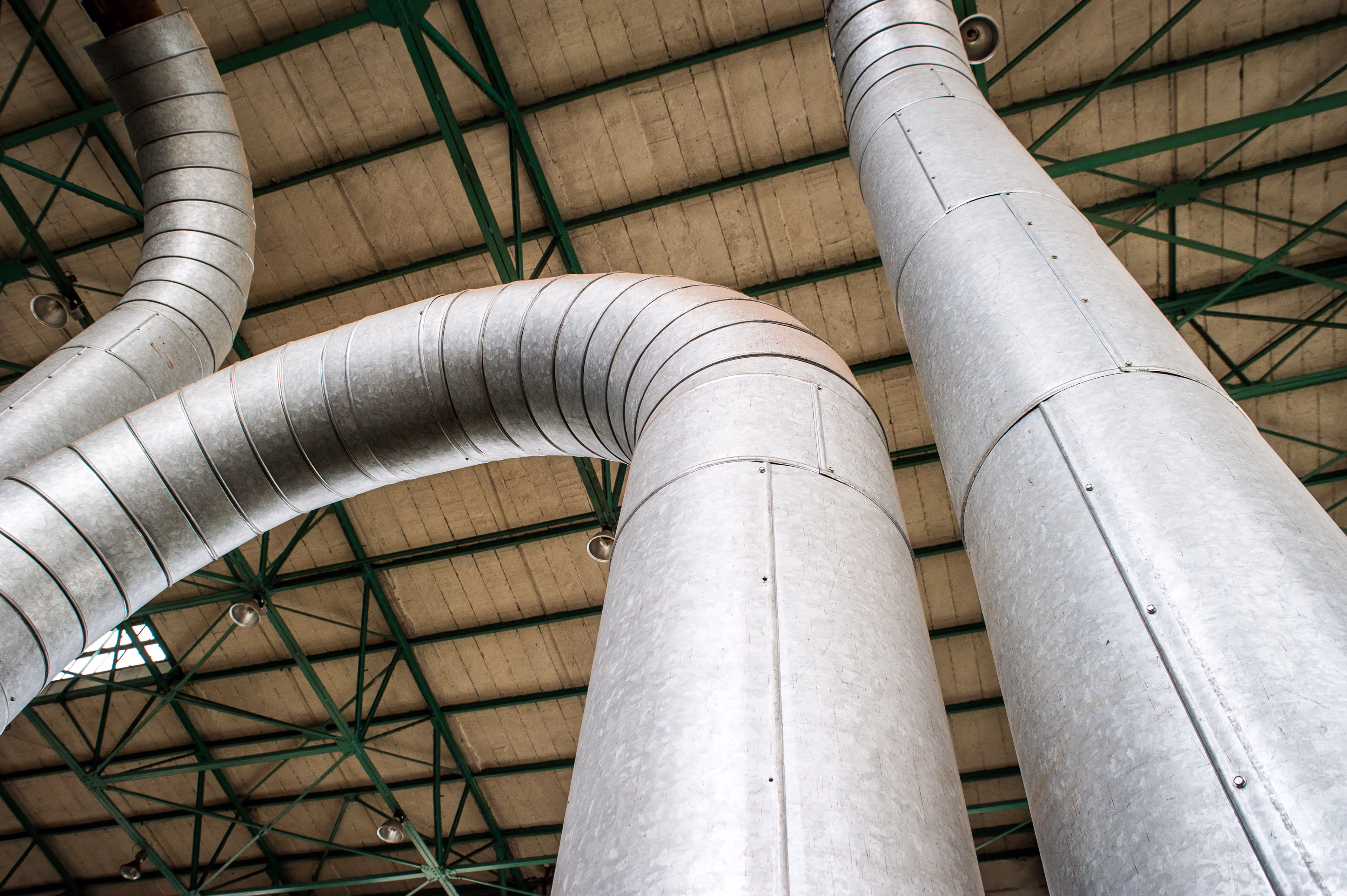 A low-angle view of a large industrial interior showing massive galvanized steel ventilation ducts. The ducts curve and extend vertically toward the ceiling, which is supported by a green metal truss framework and covered with beige acoustic panels. The surface of the ducts is textured with a metallic, slightly mottled finish and secured with visible seams and rivets. Ceiling lights hang from the beams, providing illumination. The scale and curvature of the ducts give a sense of depth and industrial complexity, emphasizing the mechanical infrastructure of the building.