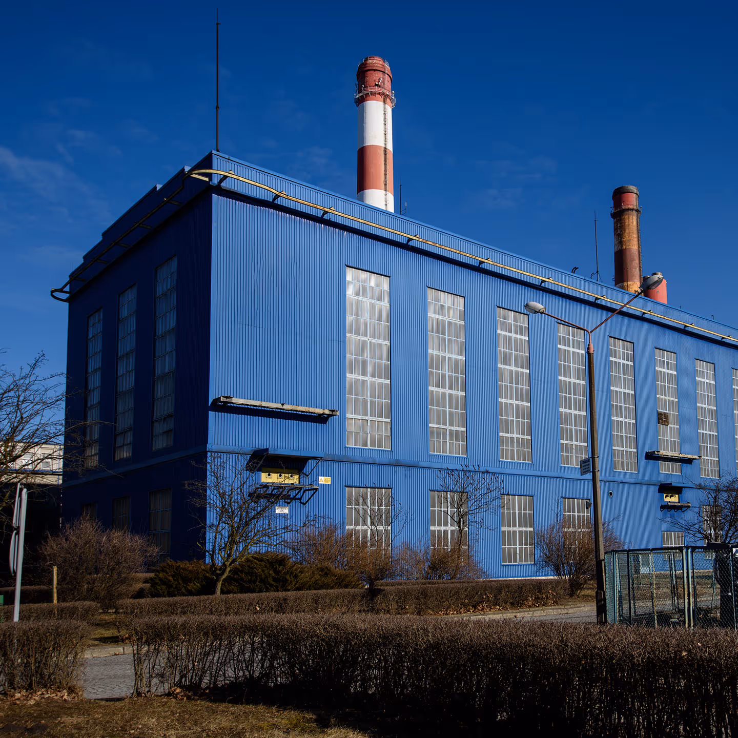 ChatGPT řekl:
A large industrial building painted in a vivid blue color with tall, narrow windows evenly spaced along its walls. The windows are translucent and arranged in vertical rows, letting in natural light. The structure has a corrugated metal exterior and a flat roof with antennas and utility fixtures on top. Behind the building, two tall smokestacks rise into the sky—one painted in red and white stripes and the other darker and weathered, indicating heavy use. In front of the building is a neatly maintained area with trimmed hedges, leafless bushes, a small fence, and a few bare trees, suggesting a late autumn or early spring season. A streetlamp stands in the foreground, and the sky is deep blue with scattered clouds.