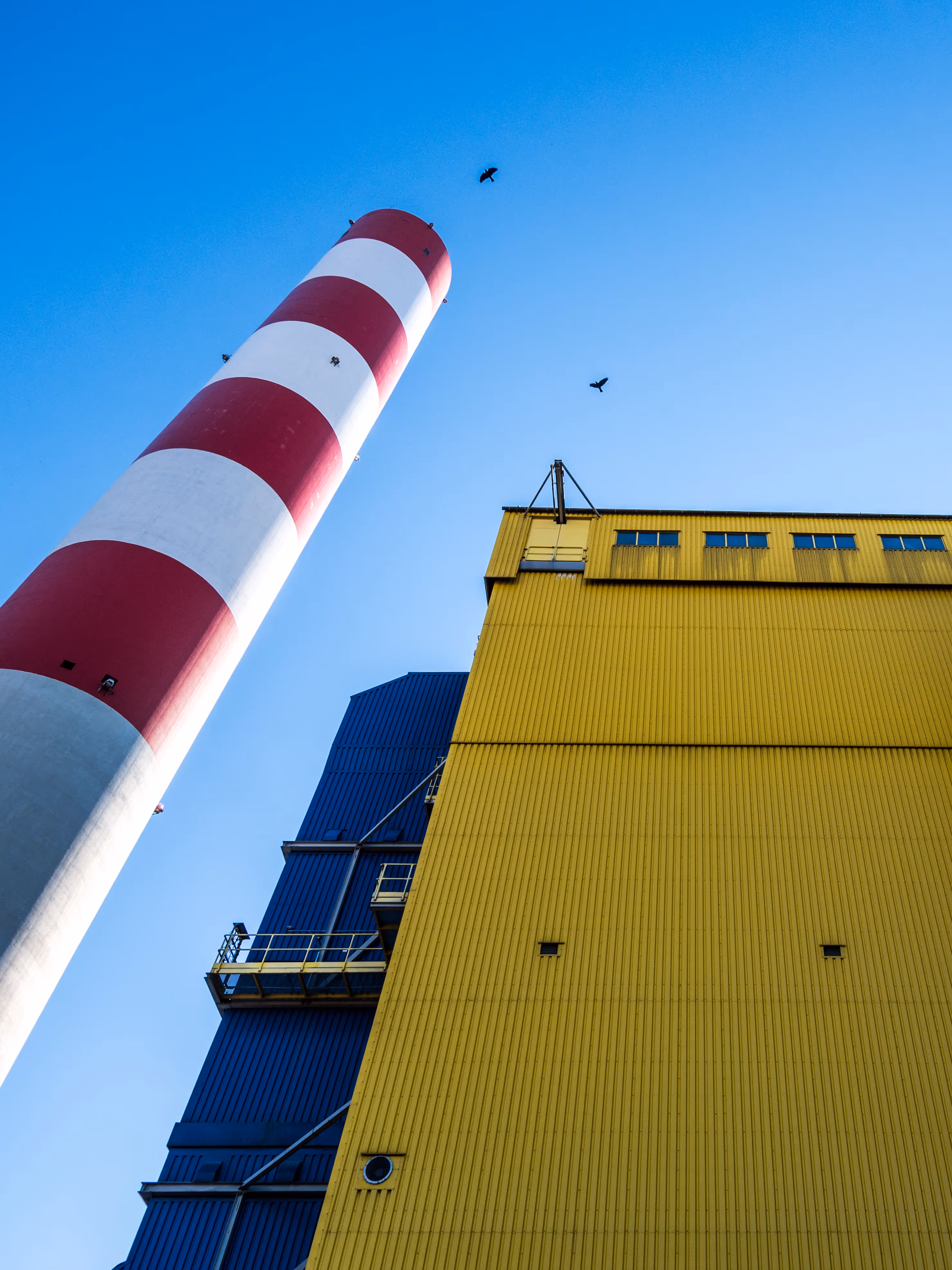 A tall industrial scene viewed from a low angle against a clear blue sky. On the left, a towering smokestack with bold red and white horizontal stripes stretches upward. To the right, a massive yellow corrugated metal building dominates the frame, its wall marked with a few small square vents and a round fixture near the base. Behind it, part of a dark blue industrial structure is visible, including a yellow-railed platform. Three birds fly high above the scene, adding a sense of scale and movement. The overall atmosphere is clean, geometric, and industrial.