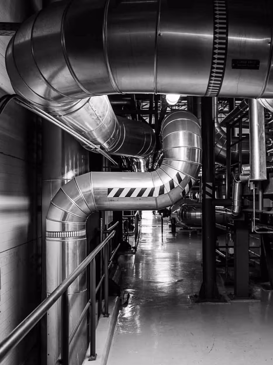 Industrial interior with large, shiny metal ventilation ducts curving and running along the ceiling and walls of a factory or utility room; the image is in black and white, highlighting the smooth texture of the pipes, the reflective surface of the clean polished floor, and the complex network of structural beams and machinery in the background.