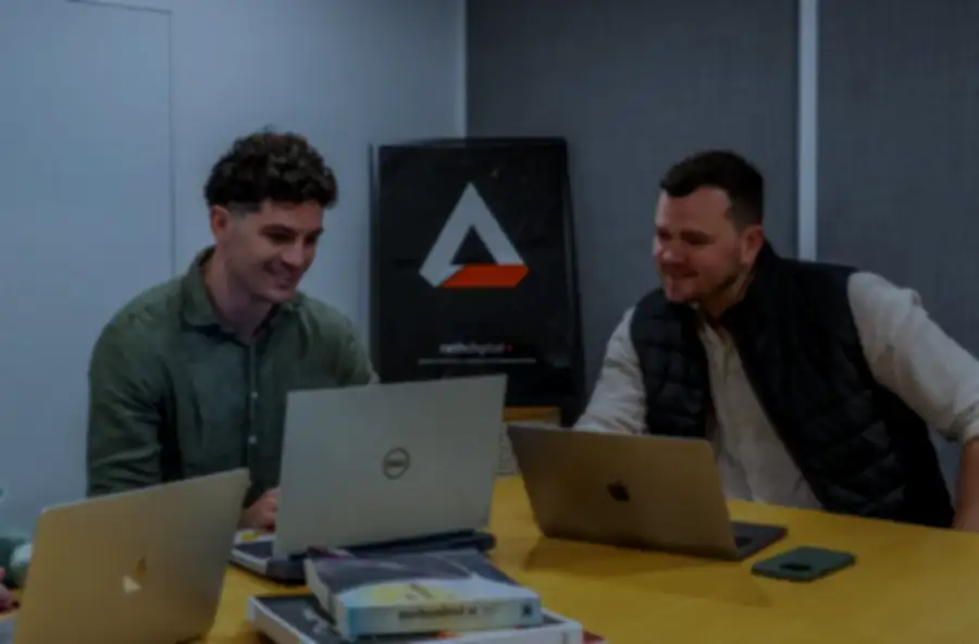 Two men sitting at a yellow table in a meeting room, each using a laptop, with a few books stacked on the table and a black and orange triangular logo on a board behind them.