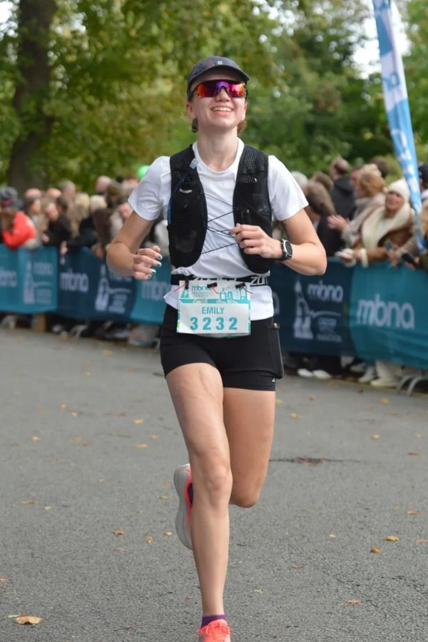 Woman in purple shirt lifting dumbbells overhead with assistance from a trainer in white shirt at the gym.