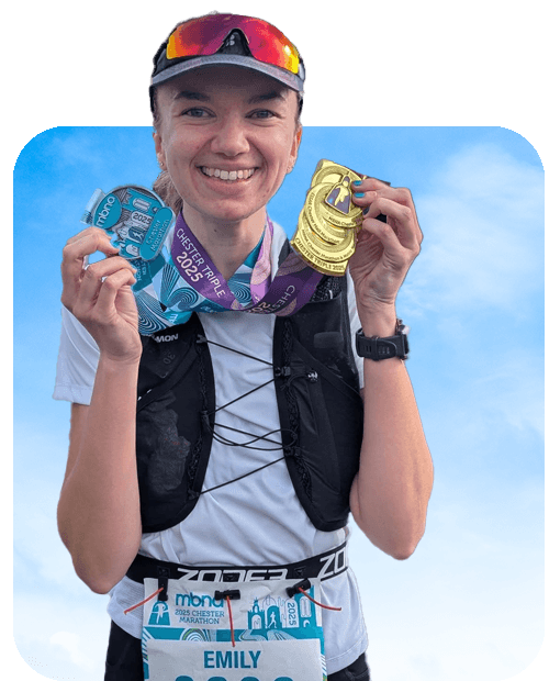 Smiling female runner named Emily wearing a white shirt, black running vest, and cap, holding two medals from the 2023 Chester Marathon against a blue sky background.