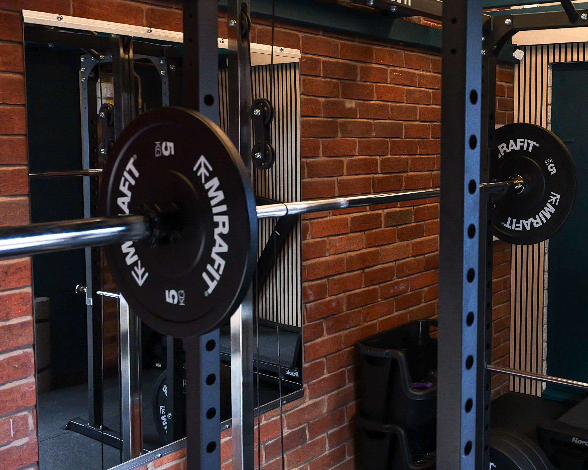 Indoor gym scene with a barbell loaded with 5 kg Mirafit weight plates mounted on a squat rack against a brick wall and a large mirror reflecting part of the equipment.