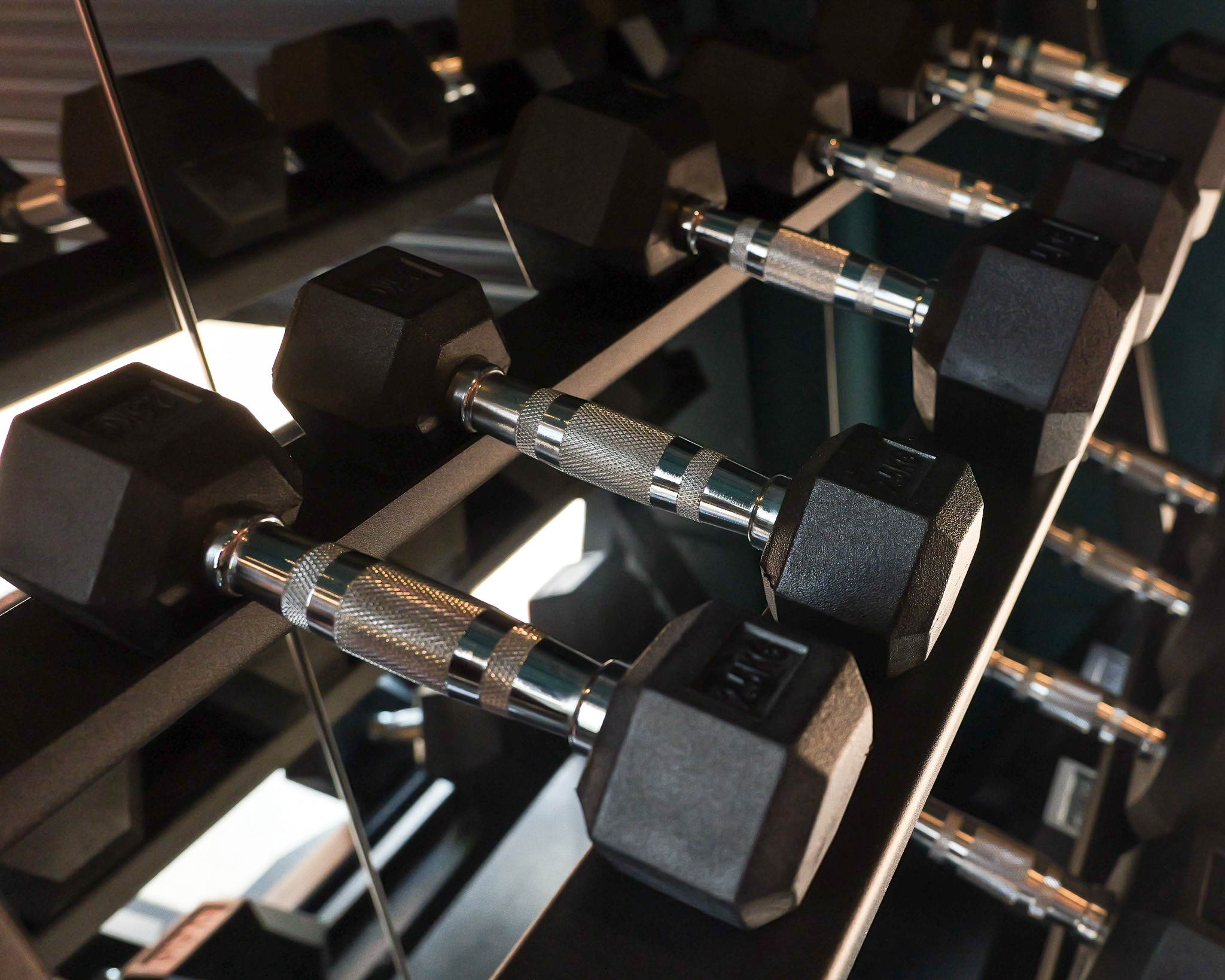 Woman in purple shirt lifting dumbbells overhead with assistance from a trainer in white shirt at the gym.